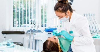 A dentist checks a woman's teeth in a dental office, with dental equipment visible in the background.